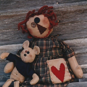 May include: A red-haired rag doll wearing a plaid dress with a heart-shaped pocket sits on a wooden bench with a brown teddy bear wearing a blue shirt.