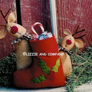 May include: Two brown felt reindeer figurines with red noses and antlers, holding a red felt stocking filled with candy and a candy cane. The stocking has a green holly design. The reindeer and stocking are in front of a wooden barn door.