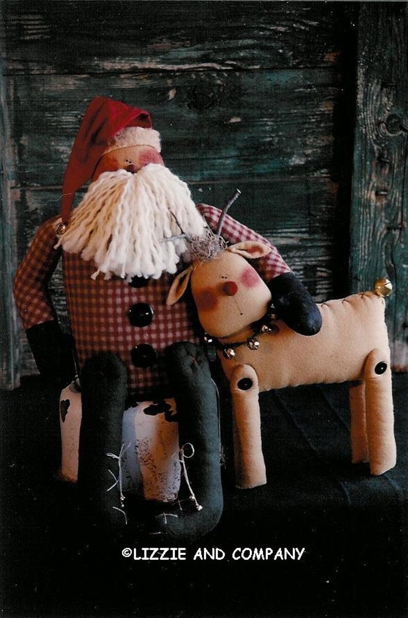 May include: A red and white plaid Santa Claus doll with a white beard sits on a wooden crate. A brown reindeer doll with black eyes and a red nose sits beside him. The dolls are handmade and have a rustic, vintage look. The image includes the text "Lizzie and Company".