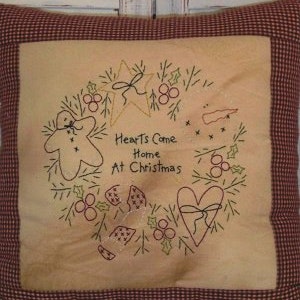 May include: A beige pillow with a red and white gingham border features an embroidered design of a wreath with a gingerbread man, a heart, a star, holly berries, and pine branches. The text "Hearts Come Home At Christmas" is embroidered in the center of the wreath.