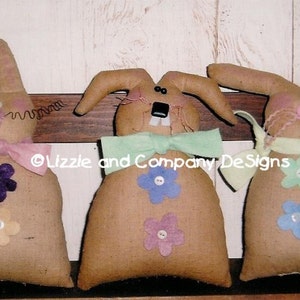 Könnte beinhalten: Drei handgefertigte Hasenpuppen mit verschiedenen Schleifen und Blumen in unterschiedlichen Farben. Die Hasen sind aus Sackleinenstoff gefertigt und haben gestickte Details. Der Text "Lizzie and Company Designs" ist im Bild sichtbar.