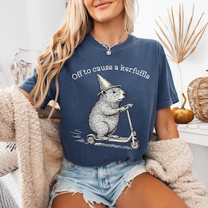 Könnte beinhalten: Marineblaues T-Shirt mit dem Schriftzug "Off to cause a kerfuffle" und einer Grafik eines Nagetiers auf einem Roller, das einen Zaubererhut trägt. Das T-Shirt hat eine lockere Passform.