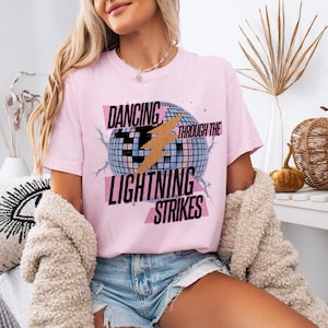 Puede incluir: Camiseta rosa claro con el texto "DANCING THROUGH THE LIGHTNING STRIKES" en negro y rosa. El diseño incluye una bola de discoteca y un rayo dorado. Parte de una colección de ropa.