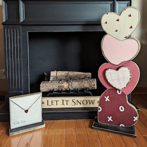 May include: A stack of four wooden hearts in shades of pink, red, and white with different patterns, including polka dots, plaid, and Xs and Os. The hearts are stacked in front of a black fireplace with a sign that reads "Let It Snow".