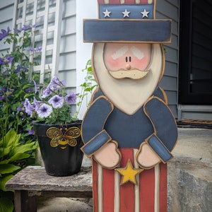 May include: A wooden Uncle Sam figure, painted in patriotic colors. The figure has a tall hat with stars, a blue coat, and red and white striped pants. A yellow star is held in the hands. The figure is standing on a wooden base.