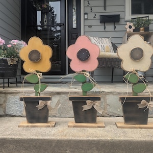 May include: Three wooden flower decorations in yellow, pink, and cream, each with a dark center and green leaves. They are in black pots with tan bows, set on a stone porch. The flowers are a charming outdoor decor item.
