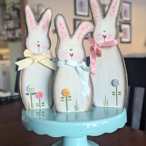May include: Three wooden bunny figurines with painted details, wearing pastel ribbon bows and standing on a light blue cake stand. Each bunny has a flower painted on its front.