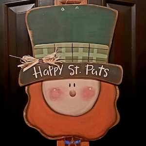 May include: A wooden door hanger shaped like a leprechaun's head with a green hat and a red beard. The sign says "Happy St. Pats".