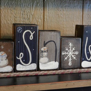 May include: Wooden blocks painted with the word "SNOW" in white paint. Each block features a different winter-themed design, including a snowman, a snowflake, and a stylized "S" and "W".