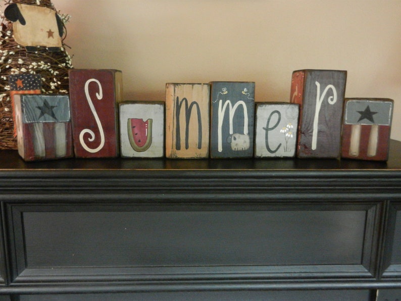以下が含まれることがあります： 各ブロックにさまざまなペイントデザインが施された、"Summer"という単語を綴る木製ブロック。ブロックは赤、青、白、茶色の色合いでペイントされています。