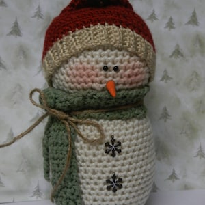 May include: A crocheted snowman with a red and beige hat, a green scarf, and brown snowflake embellishments.
