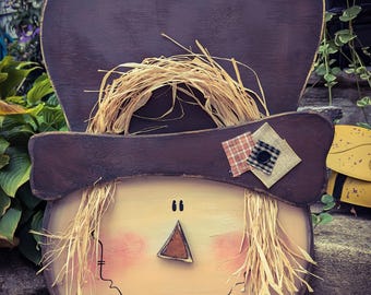 May include: A wooden scarecrow face with a brown hat, straw hair, and a painted smile. The scarecrow is decorated with a plaid and beige fabric patch.