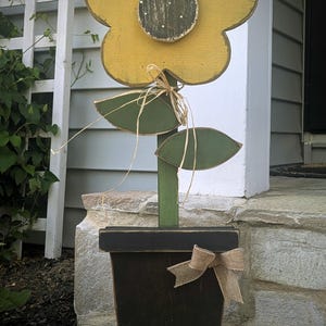 May include: A wooden sunflower decoration with a yellow flower head, green stem and leaves, and a black pot. The flower has a distressed finish, and the pot is adorned with a burlap bow. The sunflower is approximately 36 inches tall.