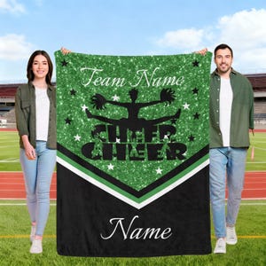 May include: A green and black blanket featuring the words "Team Name", "CHEER", and "Name". The blanket design includes a cheerleader silhouette and star motifs. Two people are holding the blanket in front of a sports field.