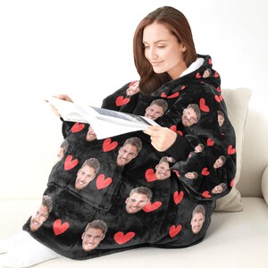 Pu&ograve; includere: Coperta nera indossabile con cappuccio, con un motivo di cuori rossi e facce sorridenti. La coperta ha un'ampia tasca frontale e fodera bianca all'interno del cappuccio. La persona sta leggendo un libro mentre indossa la coperta.