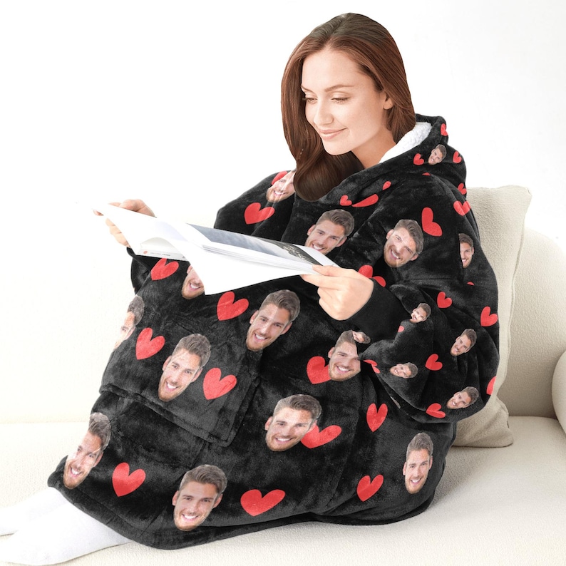 Pu&ograve; includere: Coperta nera indossabile con cappuccio, con un motivo di cuori rossi e facce sorridenti. La coperta ha un'ampia tasca frontale e fodera bianca all'interno del cappuccio. La persona sta leggendo un libro mentre indossa la coperta.