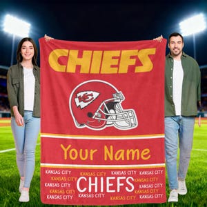 Pode incluir: Uma manta vermelha com a palavra "CHIEFS" em dourado e um gráfico de capacete de futebol. A manta também inclui o texto "Your Name" e as palavras "Kansas City" repetidas. A manta é segurada por duas pessoas.