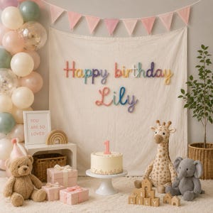 May include: A birthday party setup featuring a cream-colored fabric backdrop with the text Happy birthday Lily in colorful yarn letters. A white cake with a number one candle, plush giraffe and elephant toys, and a teddy bear are present.