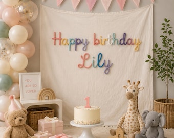 Könnte beinhalten: Ein Geburtstags-Set mit einem cremefarbenen Stoffhintergrund und dem Schriftzug Happy birthday Lily aus bunten Garnbuchstaben. Enthält eine weiße Torte mit einer Kerze in Form einer Eins, Giraffen- und Elefanten-Plüschtiere sowie einen Teddybären.