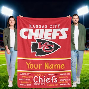 Pode incluir: Uma manta vermelha com as palavras "Kansas City Chiefs" em branco, um logotipo da equipa e espaço para um nome personalizado em amarelo. A manta tem um padrão repetido de "Kansas City Chiefs" na parte inferior.