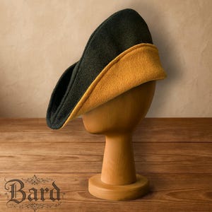 May include: A two-tone hat, with a dark green exterior and a mustard yellow interior, is displayed on a wooden head form. The hat has a curved, asymmetrical shape. The word "Bard" is visible in the lower left corner.
