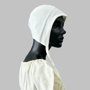 May include: A white bonnet with a brim and long ties, displayed on a black mannequin head. The bonnet is made of a textured fabric and is part of a historical costume. The mannequin is wearing a cream-colored blouse with lace trim.