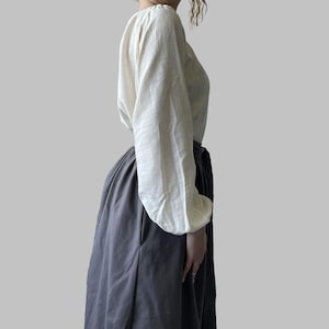 K&ouml;nnte beinhalten: Eine cremefarbene Bluse mit langen, volumin&ouml;sen &Auml;rmeln und einem Rundhalsausschnitt. Dazu wird ein langer, dunkelgrauer Rock getragen. Das Outfit besteht aus einem weichen, flie&szlig;enden Stoff und erzeugt eine schlichte, aber elegante Silhouette.