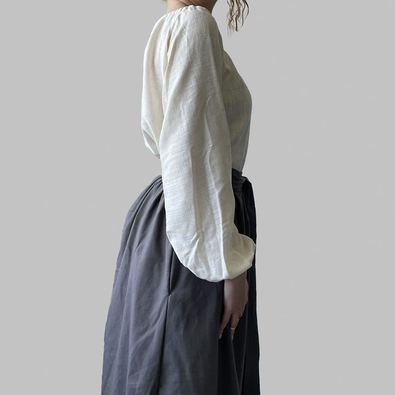 K&ouml;nnte beinhalten: Eine cremefarbene Bluse mit langen, volumin&ouml;sen &Auml;rmeln und einem Rundhalsausschnitt. Dazu wird ein langer, dunkelgrauer Rock getragen. Das Outfit besteht aus einem weichen, flie&szlig;enden Stoff und erzeugt eine schlichte, aber elegante Silhouette.
