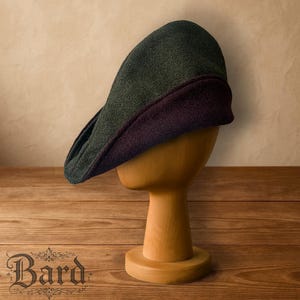 May include: A two-tone flat cap in green and burgundy, displayed on a wooden hat stand. The cap has a classic, rounded shape with a structured brim. The word "Bard" is visible in the lower left corner.