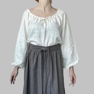 K&ouml;nnte beinhalten: Eine cremefarbene Bauernbluse mit langen, gerafften &Auml;rmeln und einem U-Ausschnitt. Die Bluse hat ein Bindedetail am Ausschnitt. Kombiniert mit einem grauen, hoch taillierten Rock mit gebundener Taille. Das Outfit ist schlicht und elegant.
