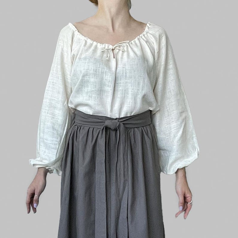 K&ouml;nnte beinhalten: Eine cremefarbene Bauernbluse mit langen, gerafften &Auml;rmeln und einem U-Ausschnitt. Die Bluse hat ein Bindedetail am Ausschnitt. Kombiniert mit einem grauen, hoch taillierten Rock mit gebundener Taille. Das Outfit ist schlicht und elegant.