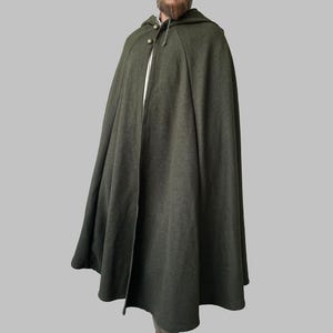 Puede incluir: Una capa con capucha verde oscuro con cierre de botón. La capa está hecha de una tela gruesa y texturizada y tiene una capucha generosa. La capa está diseñada para usarse sobre otras prendas.