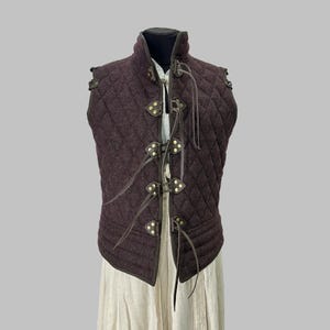 May include: A burgundy quilted vest with a diamond pattern, displayed on a mannequin. The sleeveless garment has leather detailing and brass-coloured fasteners. It features a high collar and is designed in a medieval or historical style, suitable for reenactment.