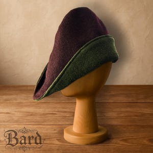 May include: A medieval-style hat with a pointed crown and a wide brim. The hat is displayed on a wooden head form. The crown is a deep burgundy colour, and the brim is a contrasting green. The hat is made of a textured fabric.