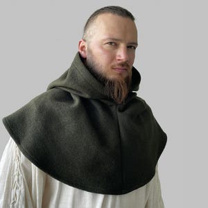 Könnte beinhalten: Ein dunkelgrüner Kapuzenmantel, vermutlich aus Wolle, mit einem breiten, abgerundeten Kragen. Die Kapuze ist am Kragen befestigt und fällt über die Schultern. Der Umhang wird über einem hellen Hemd getragen. Der Stil deutet auf mittelalterliche oder historische Kleidung hin.