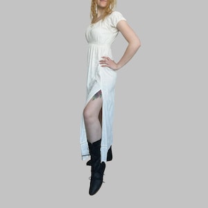 May include: A cream-colored, short-sleeved dress with a high slit. The dress features a fitted bodice and a flowing skirt. The model is wearing black lace-up boots. The dress is made of a lightweight fabric.
