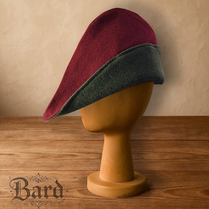 May include: A burgundy and forest green medieval hat, displayed on a wooden head form. The hat has a long, pointed crown and a contrasting band around the base. The hat is made of a felt-like material. The word "Bard" is at the bottom.