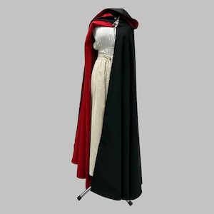 May include: A two-tone cloak, red on one side and black on the other, with a matching hood. The cloak is fastened with a decorative clasp. The garment is displayed on a mannequin, showcasing its flowing design.