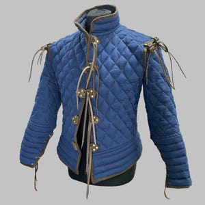 Könnte beinhalten: Gesteppte blaue Jacke mit braunen Zierleisten und Lederschnüren. Die Jacke hat einen hohen Kragen und eine Rautensteppung. Die Ärmel haben braune Lederschnüre an den Manschetten. Für historische Reenactments oder Kostüme.