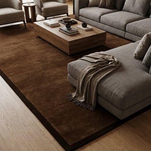May include: A large, rectangular, solid dark brown area rug with a smooth, low-pile texture. The rug is positioned on a light-toned wooden floor, extending underneath a wooden coffee table and a gray sectional sofa with a draped throw blanket.