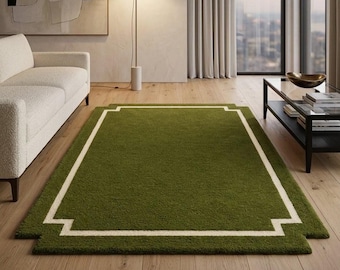 Puede incluir: Una alfombra rectangular con pelo de color verde oliva intenso y un borde en contraste de color crema. El borde forma un marco geométrico con esquinas recortadas, creando un diseño limpio y estructurado sobre un suelo de madera clara.