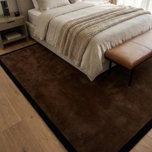 May include: A rectangular area rug featuring a deep brown center with a solid black border. The rug is positioned on a light-colored wooden floor, extending partially underneath a bed frame. The surface of the rug appears soft and uniform in texture.