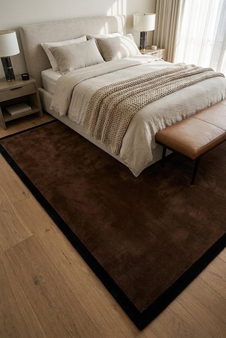 May include: A rectangular area rug featuring a deep brown center with a solid black border. The rug is positioned on a light-colored wooden floor, extending partially underneath a bed frame. The surface of the rug appears soft and uniform in texture.