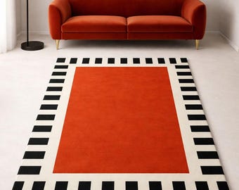 May include: A rectangular area rug with a bold design. The rug features a vibrant orange center, framed by a white border and a repeating pattern of black rectangles. The rug is placed on a light-colored floor.
