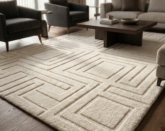 May include: A cream-colored area rug with a geometric pattern of squares and lines. The rug is placed on a dark wood floor in a living room setting. The room also features dark gray armchairs and a dark brown coffee table.