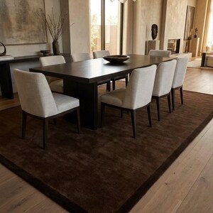 May include: A rectangular dark brown wooden dining table with six upholstered chairs in a light beige fabric. The table features a central dark brown bowl and is positioned on a large dark brown area rug. A sculptural light fixture hangs above.