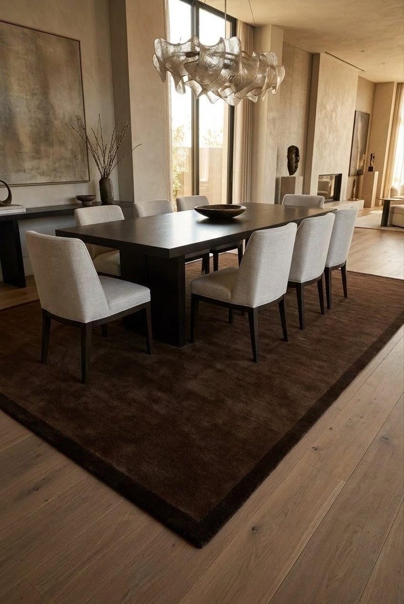 May include: A rectangular dark brown wooden dining table with six upholstered chairs in a light beige fabric. The table features a central dark brown bowl and is positioned on a large dark brown area rug. A sculptural light fixture hangs above.
