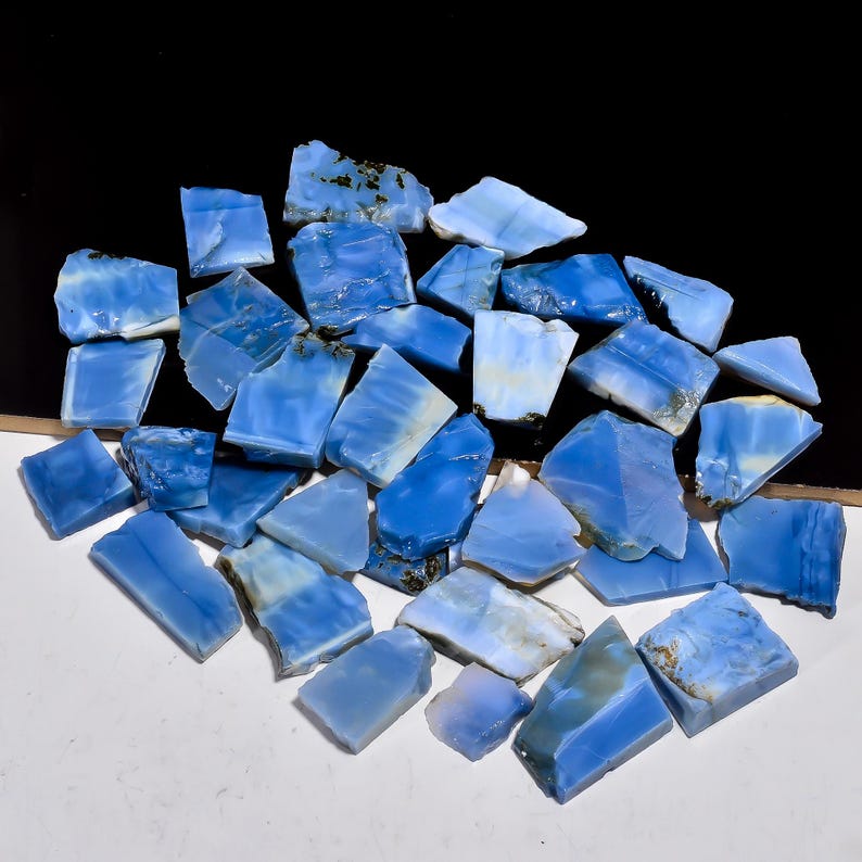 May include: A collection of numerous irregular, flat pieces of blue opal gemstone. Each fragment displays varying shades of light to medium blue with occasional white streaks and small patches of dark brown mineral inclusions on the edges.