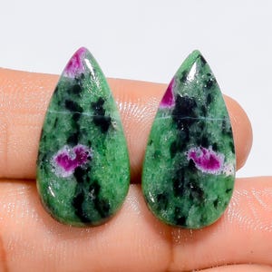 Könnte beinhalten: Zwei polierte, tropfenförmige Edelsteine. Die Steine weisen eine leuchtend grüne Grundfarbe mit schwarzen Einschlüssen und Flecken in tiefem Rosa und Magenta auf. Die Edelsteine sind vor einem neutralen Hintergrund platziert.