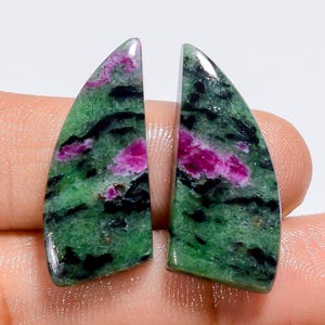 May include: Two polished ruby zoisite gemstones. The stones have a green base color with black veining and patches of vibrant pink ruby. The gemstones are cut into a curved, elongated shape, and are displayed against a neutral background.